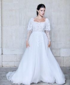 a woman standing in front of a wall wearing a white wedding dress with long sleeves