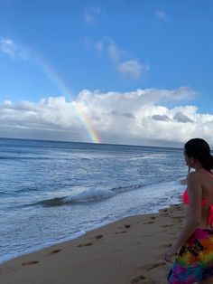 a woman standing on top of a sandy beach next to the ocean with a rainbow in the sky