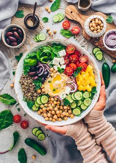 a person holding a bowl filled with vegetables and chickpeas on top of a table