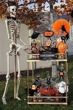 a skeleton sitting on top of a table filled with halloween decorations and candies in front of a fence