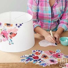 a woman sitting at a table painting flowers on a lamp shade with watercolors