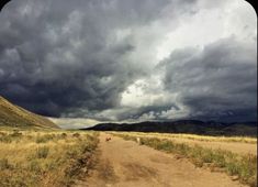 a dirt road in the middle of a field with storm clouds above it and grass on both sides