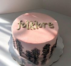 a pink and black cake with gold lettering on it sitting on a white table next to a window