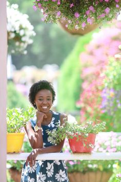 a woman standing in front of potted plants and smiling at the camera with her hand on her chin