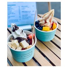 two cups filled with ice cream, fruit and chocolate on top of a wooden table