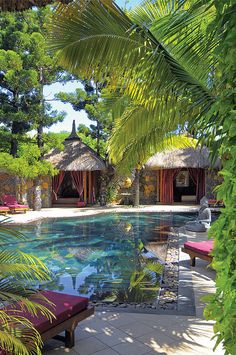 an outdoor swimming pool surrounded by palm trees and lawn chairs with thatched huts in the background