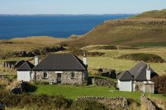 an old stone house sits on the side of a grassy hill next to some water