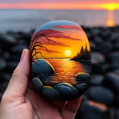 a hand holding up a painted rock near the ocean at sunset with trees on it