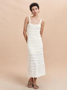 a woman standing in front of a beige background wearing a white dress and sandals with her hands on her hips