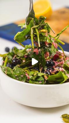 a white bowl filled with greens and blueberries on top of a table next to lemon wedges