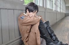 a young man sitting on the ground next to a wall wearing black boots and a brown jacket