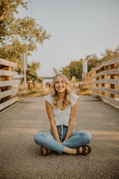 a woman sitting on the ground with her legs crossed and smiling while she sits in front of a wooden fence