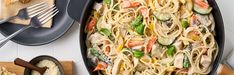 a skillet filled with pasta and vegetables on top of a table next to utensils