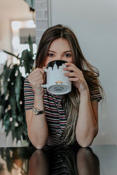 a woman sitting at a table holding a coffee cup in front of her face and looking into the camera