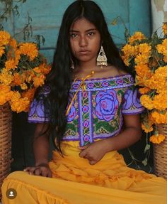 a young woman sitting in front of flowers wearing an embroidered top and yellow skirt with tassels