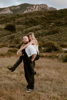 a man carrying a woman on his back in an open field with mountains in the background