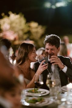 a man and woman sitting at a dinner table with wine glasses in their hands, smiling