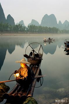 a man sitting on top of a bamboo raft next to water with mountains in the background