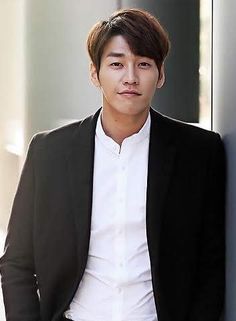 a young man leaning against a wall wearing a suit and white shirt with his hands in his pockets