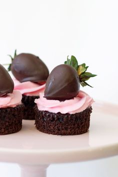 three chocolate cupcakes with pink frosting and strawberries on top sitting on a white cake plate