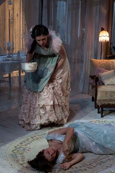 two women dressed in period clothing are laying on the floor and one is holding a bowl