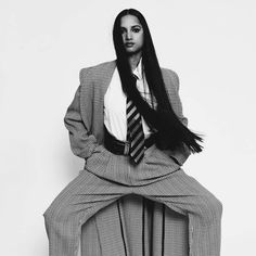 a woman with long black hair sitting on a chair wearing a suit and striped tie