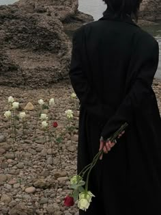 a person in a long coat holding flowers near the water with rocks and grass behind them