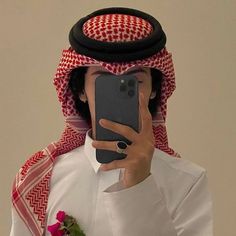 a man in an arab outfit taking a selfie with his cell phone while wearing a red and white headdress