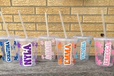 four personalized tumblers with straws are lined up on a wooden table in front of a brick wall