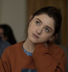 a woman sitting down with her hand on her chin and looking at the camera while wearing an orange sweater