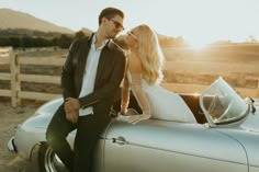 a man standing next to a woman sitting on top of a silver car in front of a wooden fence