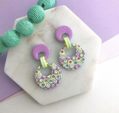 a pair of purple and green earrings sitting on top of a marble block next to yarn