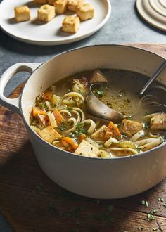 a pot filled with soup and vegetables on top of a wooden table next to plates