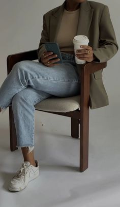 a woman sitting in a chair holding a cup of coffee and looking at her cell phone