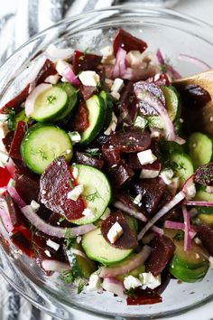 a glass bowl filled with cucumbers, onions and feta cheese next to a wooden spoon