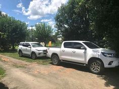 two white trucks parked next to each other on a dirt road near trees and bushes