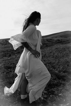 black and white photograph of woman in long dress standing on grassy field with wind blowing her hair