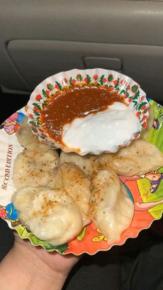 a hand holding a paper plate with food on it and sauce in the bowl next to it