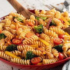 a red bowl filled with pasta and veggies next to a wooden spatula