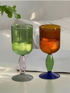 two wine glasses sitting next to each other on top of a white table with a plant in the background