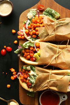 three wraps filled with vegetables and garnishes on a cutting board next to dipping sauce