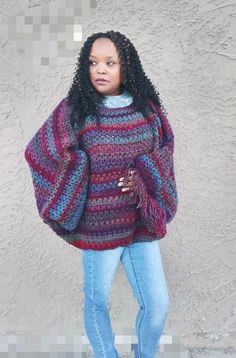 a woman standing in front of a cement wall wearing a colorful sweater and blue jeans