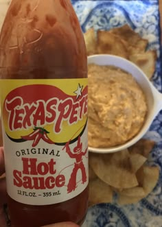 a hand holding a bottle of hot sauce next to some chips and a bowl of dip