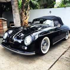 an old black car is parked in front of a house with palm trees and a white wall