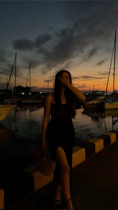 a woman in a black dress is standing near the water at sunset with boats behind her