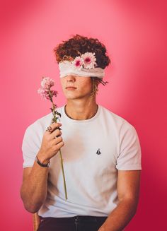 a young man with flowers in his hair holding a flower behind his head while wearing a blindfold