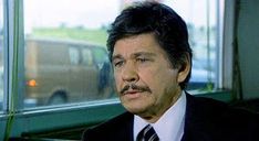 a man wearing a suit and tie sitting in front of a window with a van behind him