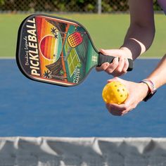 a person holding a tennis racket and ball in their hand on a court with blue water behind them