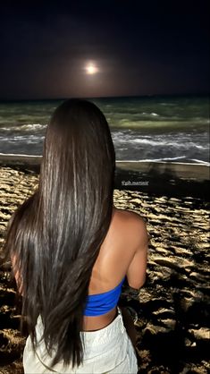 a woman sitting on top of a sandy beach under a full moon at the ocean