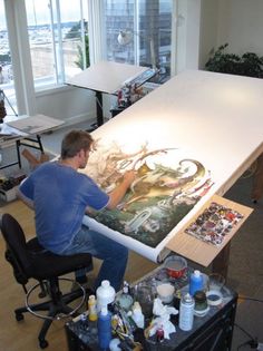 a man sitting in front of a large painting on a white table next to a window